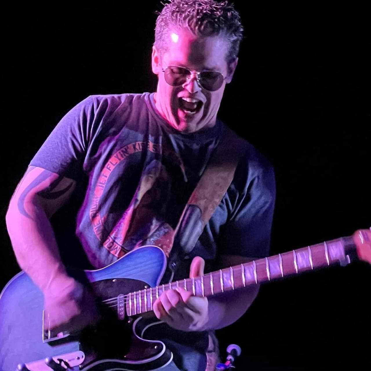 AJ Cronk playing a blue electric guitar onstage under purple lighting