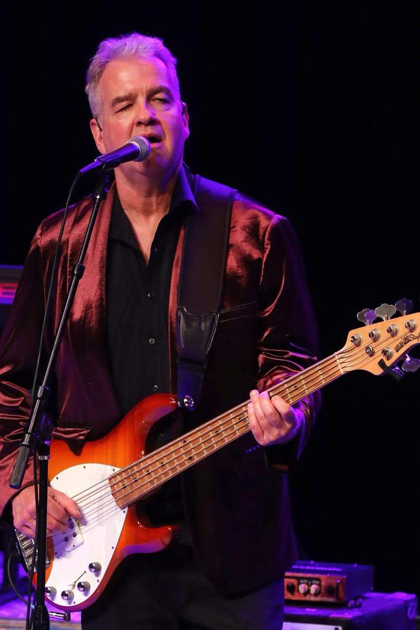 Paul Greenlease singing into a microphone while playing bass guitar onstage